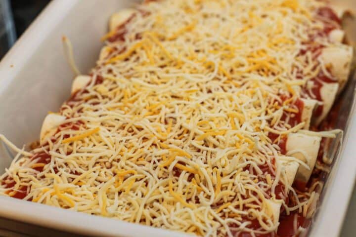 A casserole dish of enchiladas topped with cheese and red sauce before baking. 