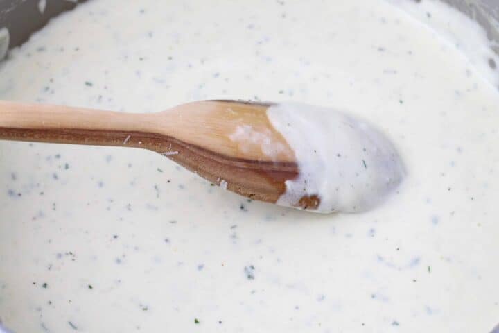 Alfredo sauce being finished in a saucepan. Image shows the sauce coating the back of a wooden spoon to show the finished consistency. 