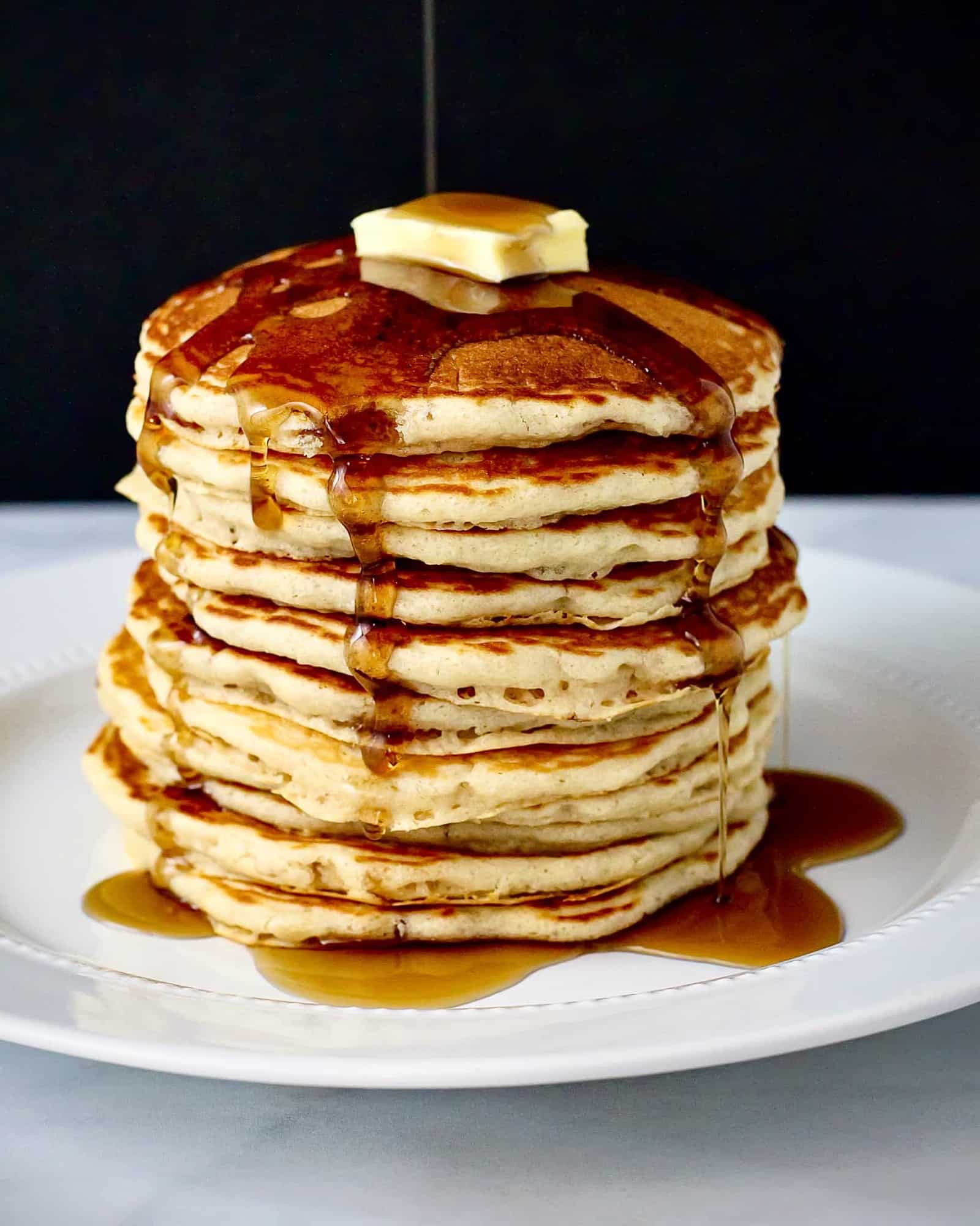 Side view of a tall stack of pancakes drizzled with maple syrup set against a black background.