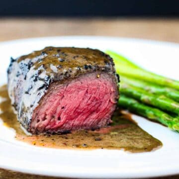 Side view of a steak topped with au poivre sauce and sliced to reveal a rosy pink interior.