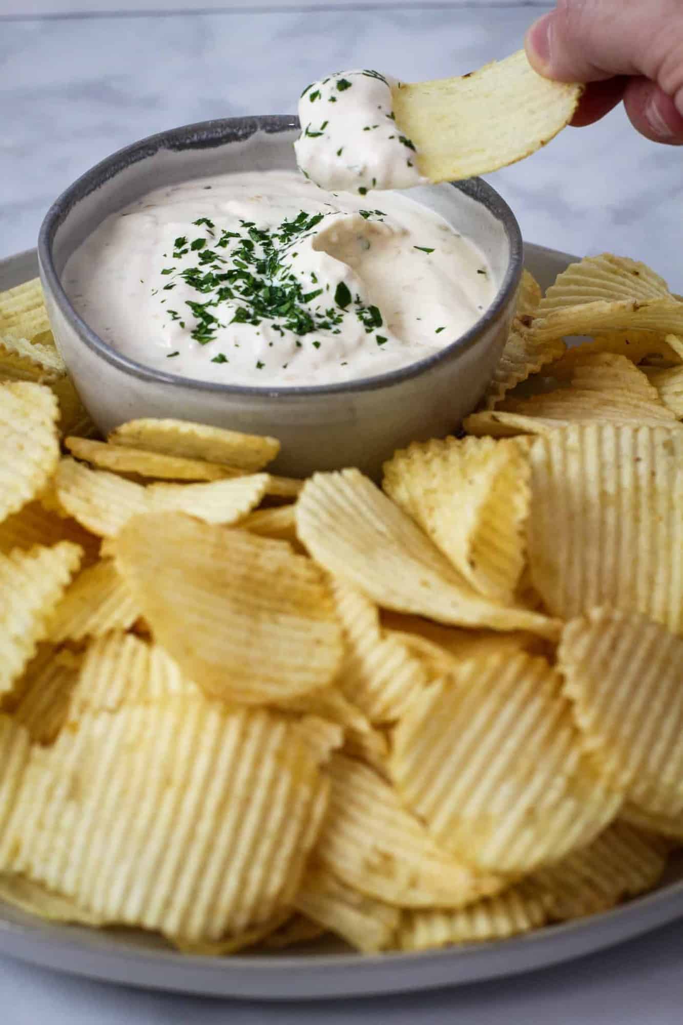The onion dip is shown served on a platter with potato chips with someone taking a scoop with a chip.