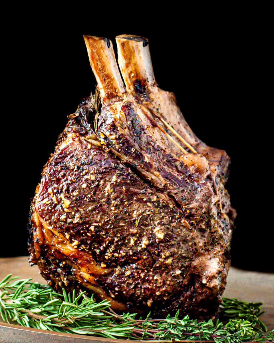 A roasted prime rib standing on the fat end with the bones pointing up, surrounded by fresh rosemary and thyme on a serving platter.