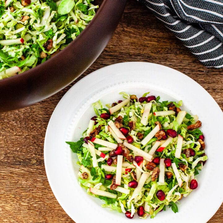 A serving of Brussels sprouts and kale salad on a plate, next to a serving bowl of salad.