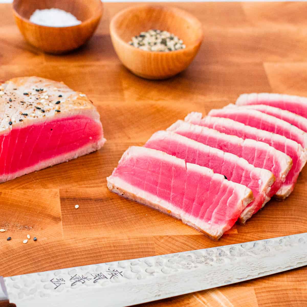 A seared tuna steak shown sliced on a large wooden cutting board with a knife in the foreground.