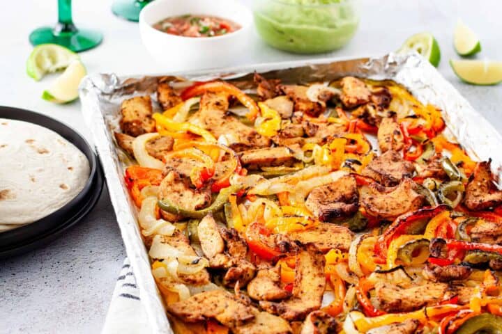 A table spread with a sheet pan of roasted chicken and vegetables served with tortillas, guacamole, pico de gallo, and lime wedges.