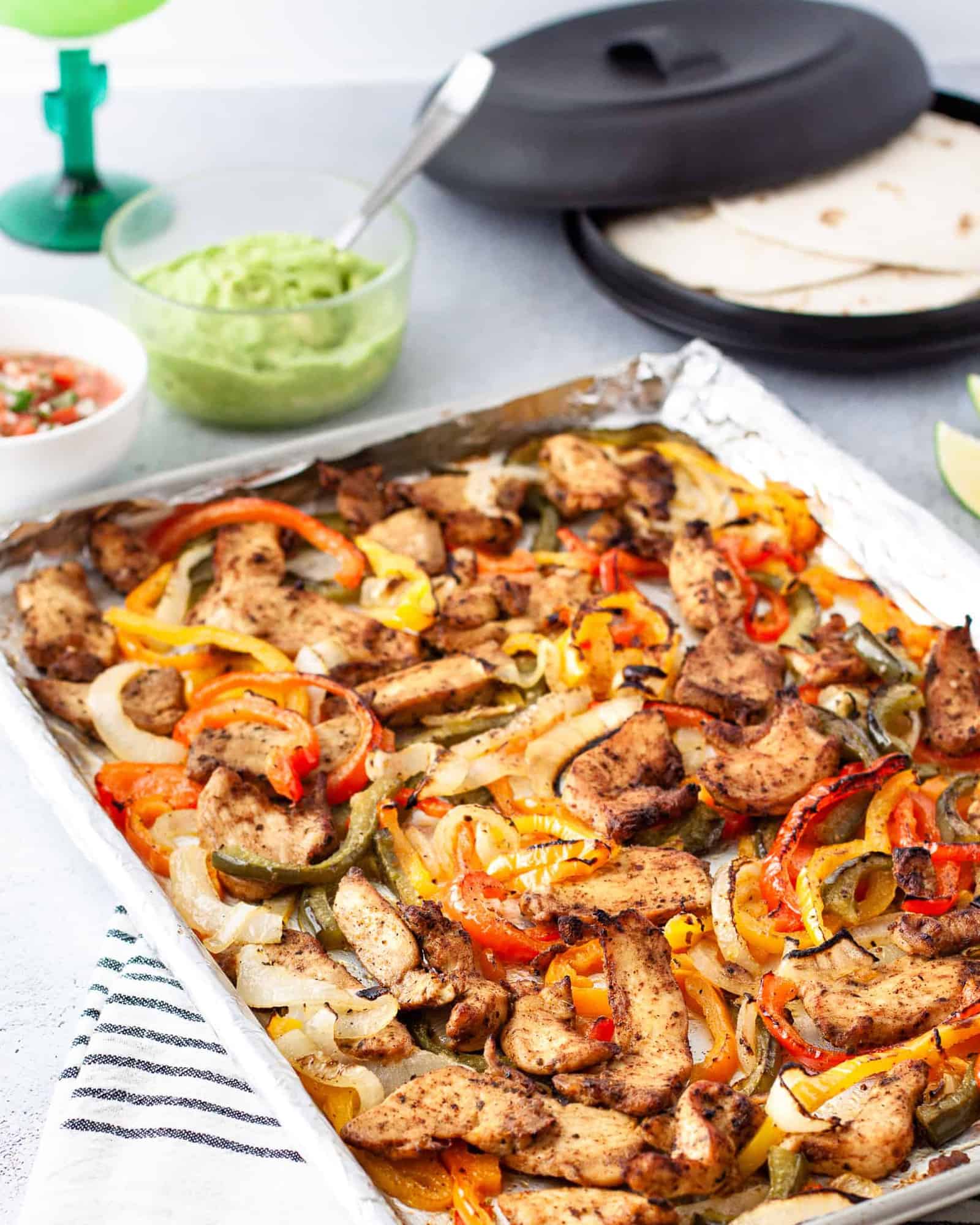 A sheet pan of chicken fajita meat and veggies on a table with tortillas and toppings. 