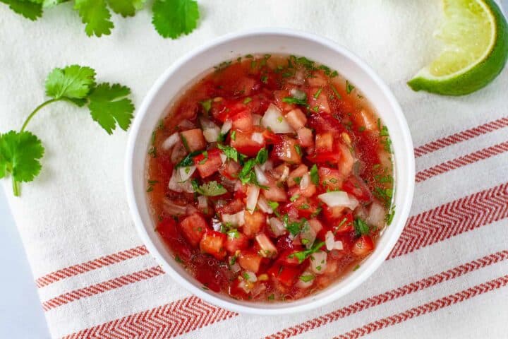 Pico de gallo salsa served in a bowl pictured on a cloth napkin with lime wedges and cilantro