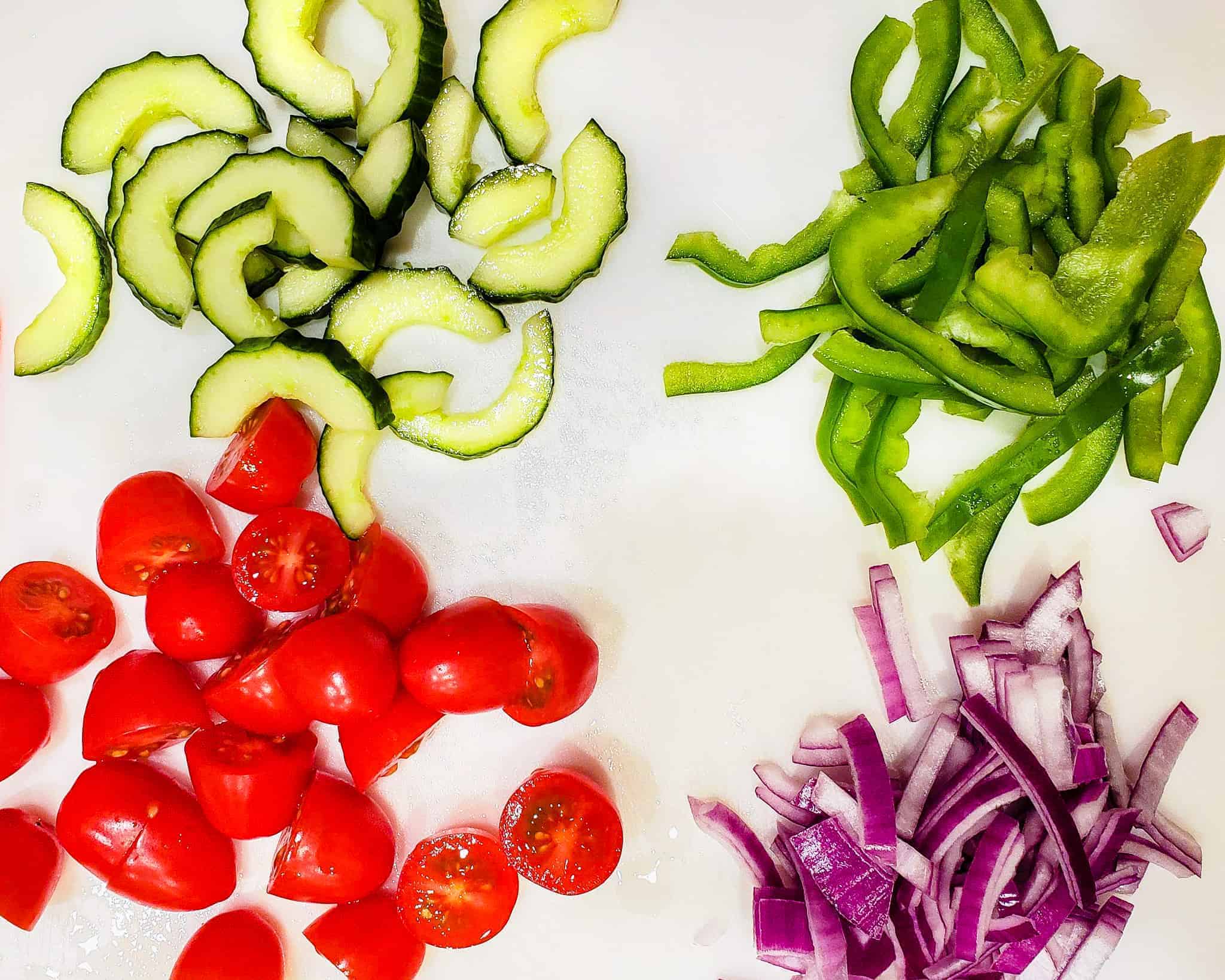 Sliced cucumber, green pepper, tomatoes, and red onion shown on a cutting board.