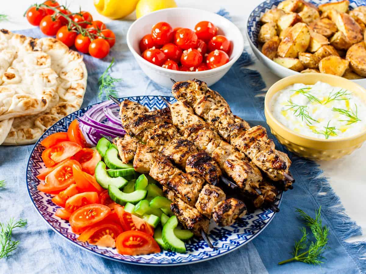 A table with a platter of chicken souvlaki and vegetables with a bowl of tzatziki dipping sauce. 