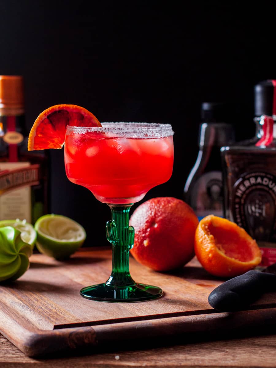 Margarita served in a sugar rimmed glass shown on cutting board with cut oranges and lime.