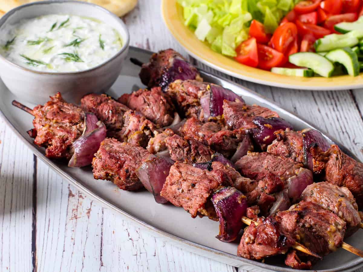 Greek yogurt marinated lamb kebabs shown served on a platter with a bowl of tzatziki sauce and fresh vegetables.