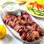Grilled lamb kebabs served with warm pita bread, a bowl of fresh tzatziki sauce, and fresh tomatoes and cucumbers.