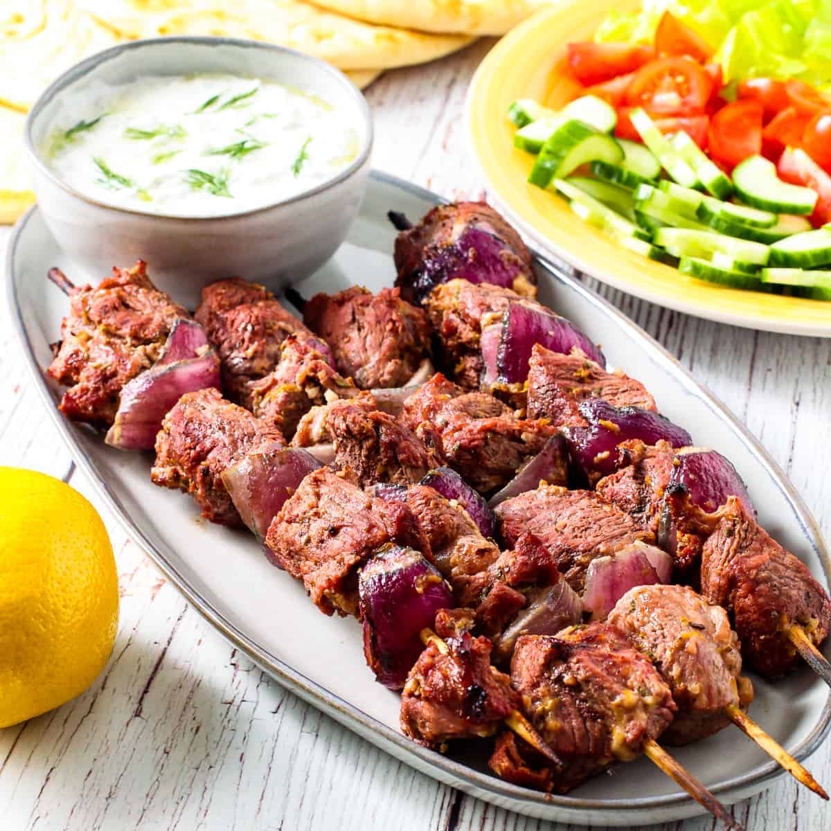 Greek yogurt marinated lamb kebabs shown served on a platter with a bowl of tzatziki sauce,warm pita bread, and a platter of fresh vegetables.