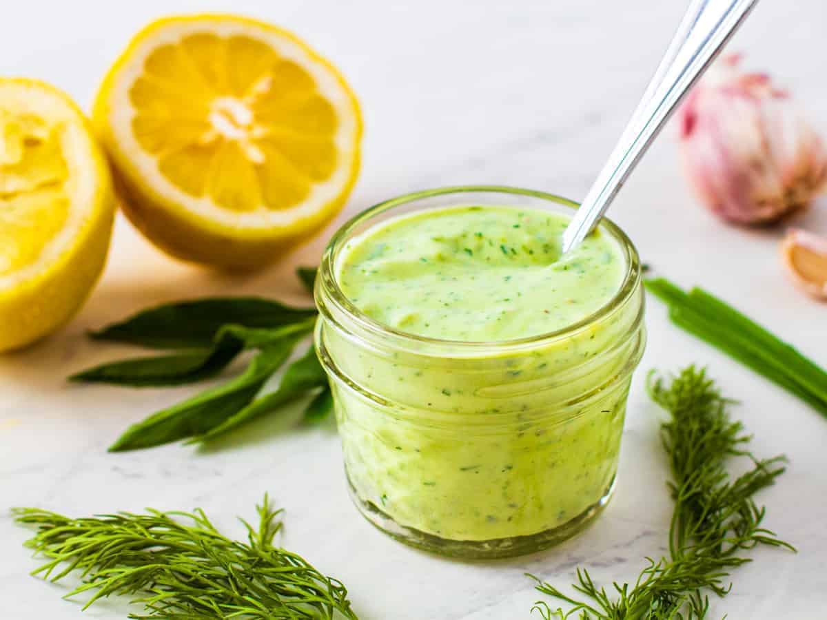 Avocado herb dressing in a jar with a spoon surrounded by fresh herbs and lemons.