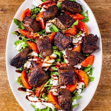 Overhead image of the steak kabobs served on an oval platter atop a salad of baby arugula, heirloom tomatoes, and burrata cheese, with a drizzle of balsamic glaze.