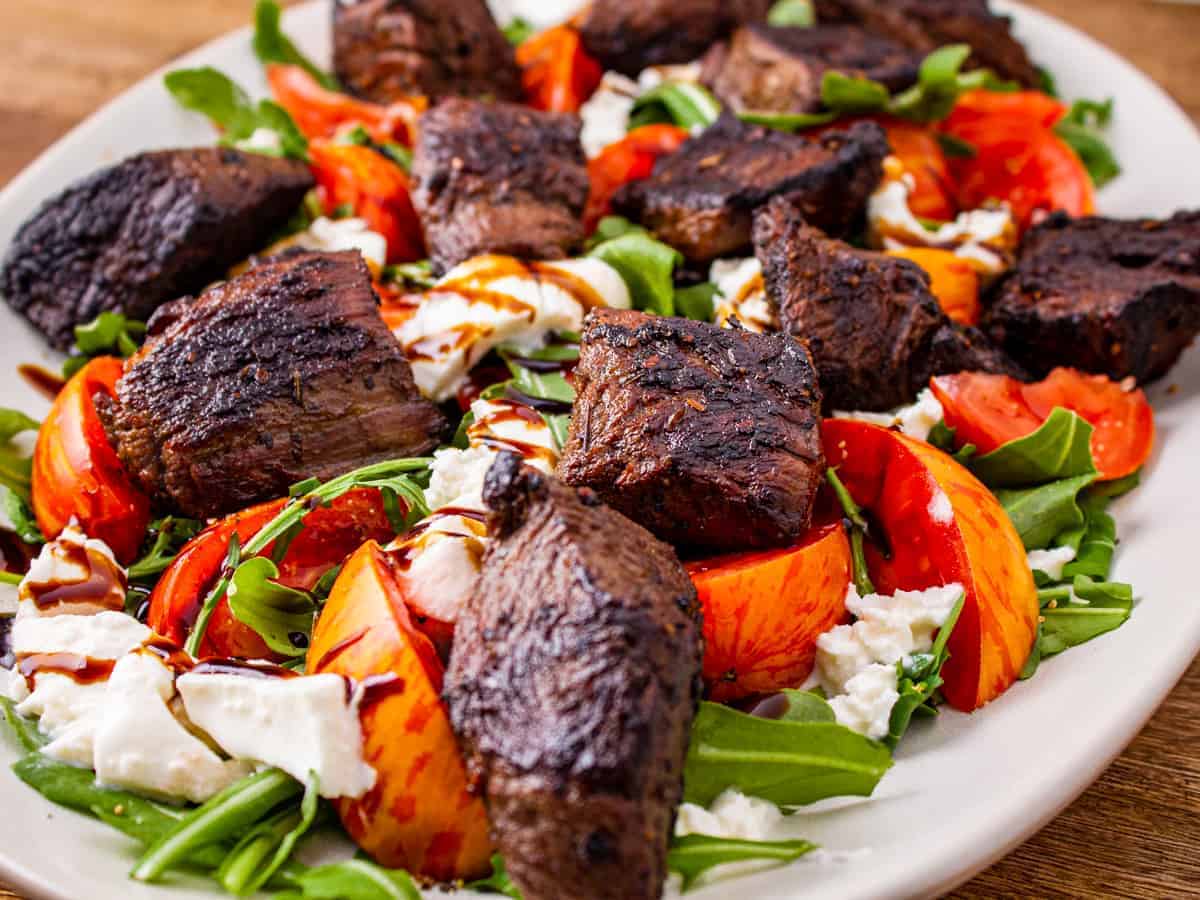 Steak Kabobs served on a platter atop a salad of baby arugula, heirloom tomatoes, and burrata cheese and topped with a drizzle of balsamic glaze.