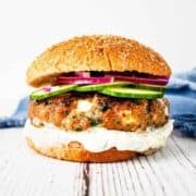 Close up image of a spinach and feta turkey burger on a wheat bun with a thick spread of tzatziki sauce and topped with sliced cucumber and red onion.