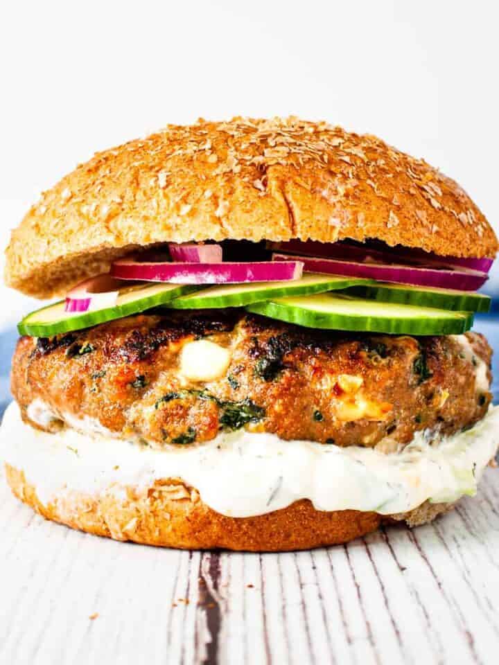 Close up image of a spinach and feta turkey burger on a wheat bun with a thick spread of tzatziki sauce and topped with sliced cucumber and red onion.