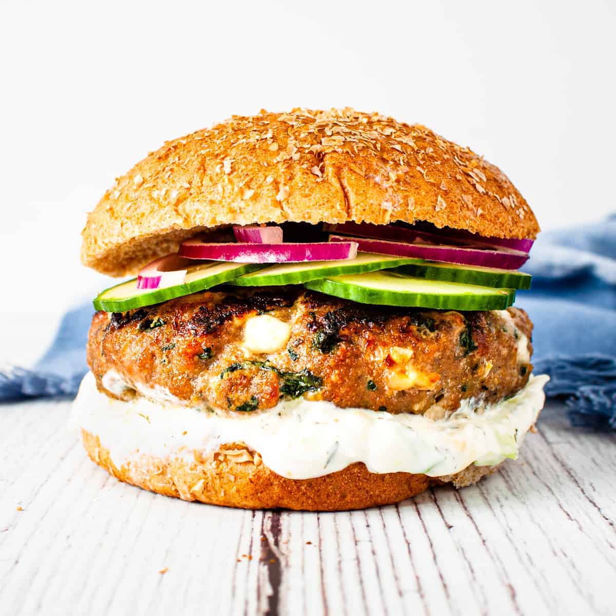 Close up image of a spinach and feta turkey burger on a wheat bun with a thick spread of tzatziki sauce and topped with sliced cucumber and red onion. 