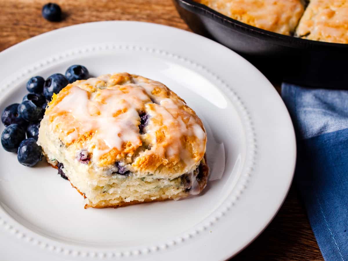 Close up view of a glazed blueberry buttermilk biscuit served on a white dish with fresh blueberries.