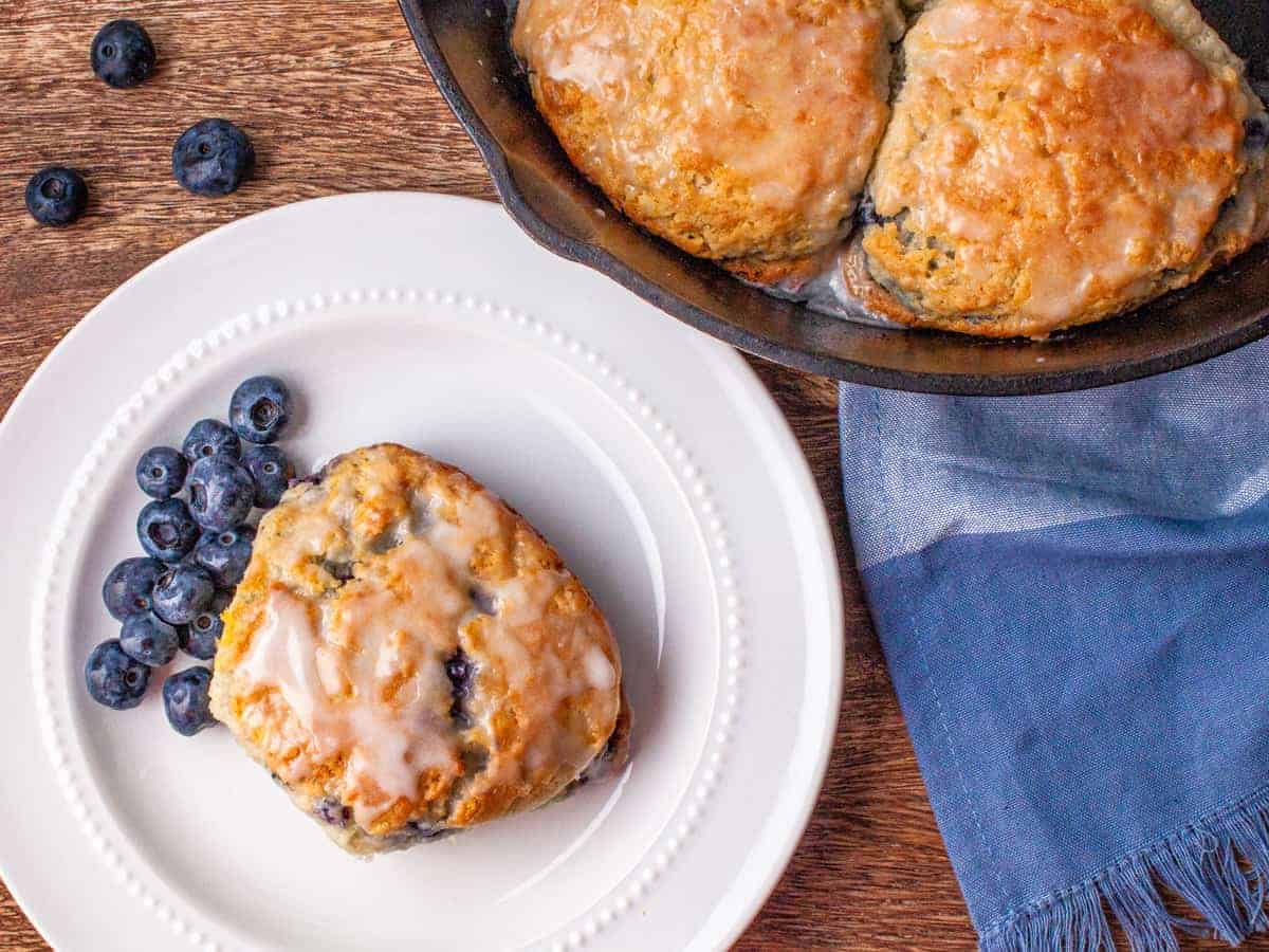 Overhead view of a biscuit served on a white dish next to a cast iron skillet of blueberry buttermilk biscuits.