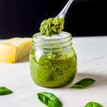 Image of a spoon dipping into a jar of homemade pesto on a white countertop against a black background.