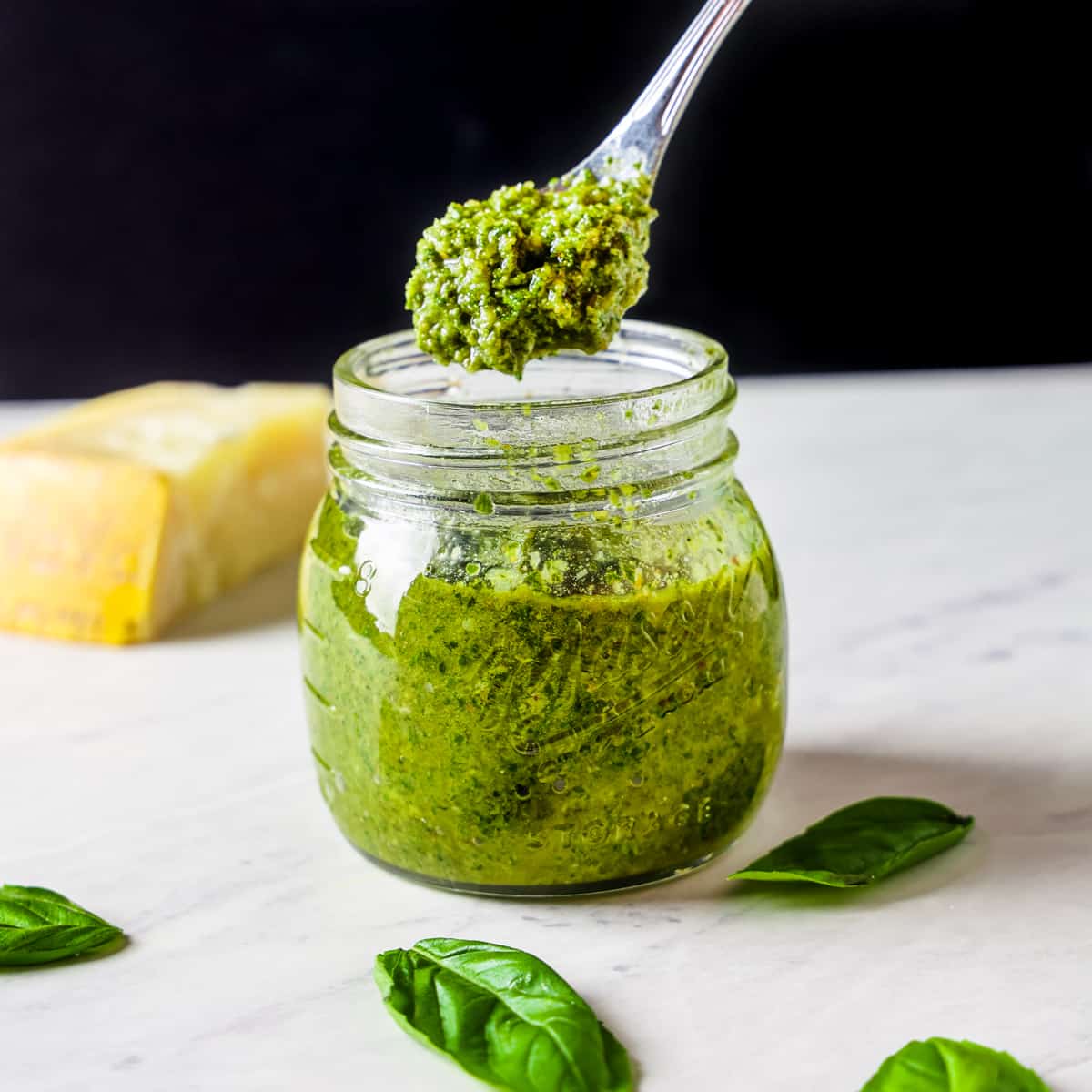 Image showing a spoon dipping into a jar of homemade pesto sauce set on a white countertop against a black background.