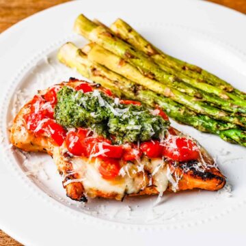Side angle view of a grilled chicken breast topped with mozzarella cheese, chopped cherry tomatoes, and basil pesto served with a side of grilled asparagus.
