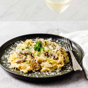A plate of creamy mushroom pasta is shown set on a white countertop garnished with grated Parmesan cheese and parsley.