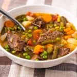 Side angle view of a bowl of vegetable beef soup served in a bowl on a plaid tablecloth.