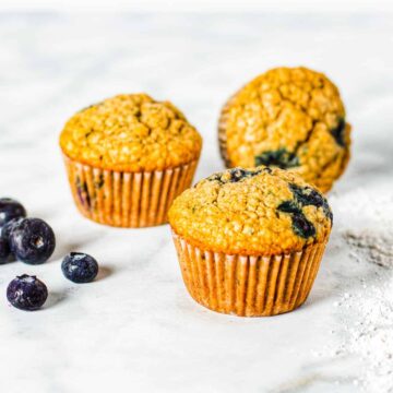 Three blueberry muffins shown on a countertop with fresh blueberries.