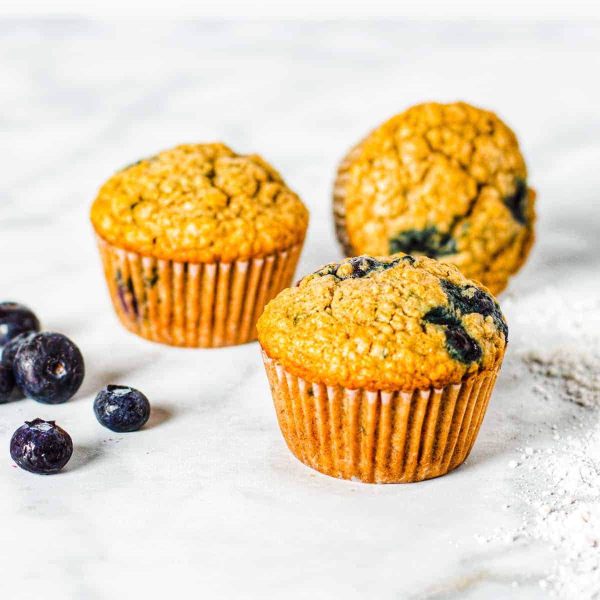 Three blueberry oatmeal muffins shown on a countertop with fresh blueberries.