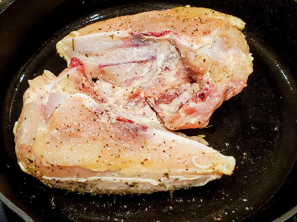 Bone-in, skin on chicken breasts shown being seared in a cast iron skillet.