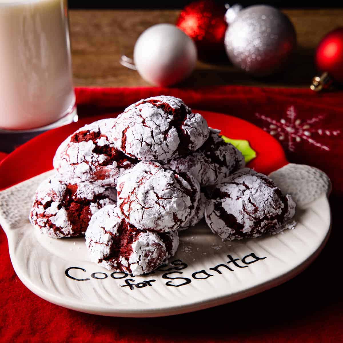 Plate of red velvet crinkle cookies shown on a table with a red placemat and surrounded by Christmas ornaments. 