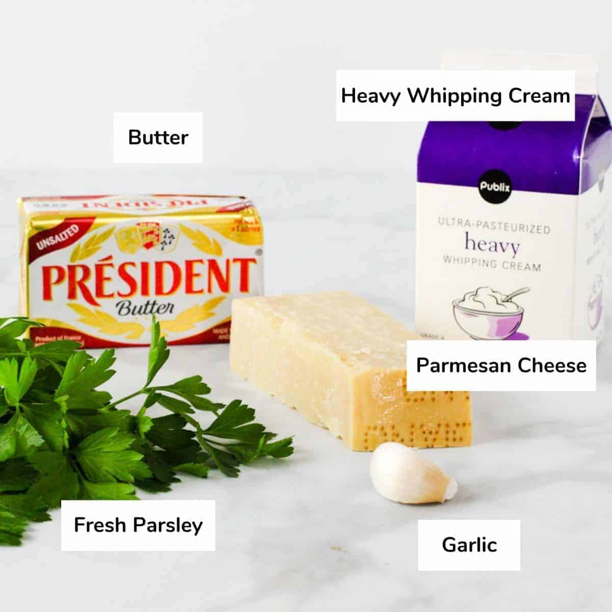 The ingredients for alfredo sauce; butter, heavy whipping cream, Parmesan cheese, garlic, and fresh parsley, are shown set on a counter.