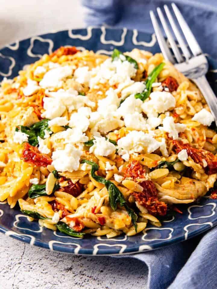 Side angle view of a plate of chicken and orzo with sun-dried tomatoes and spinach topped with crumbled feta cheese.