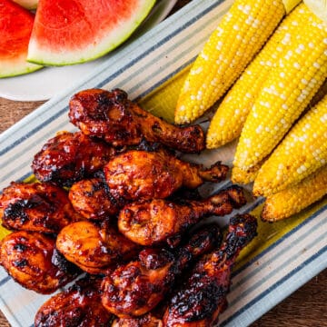 A platter of grilled chicken legs served with corn and watermelon.