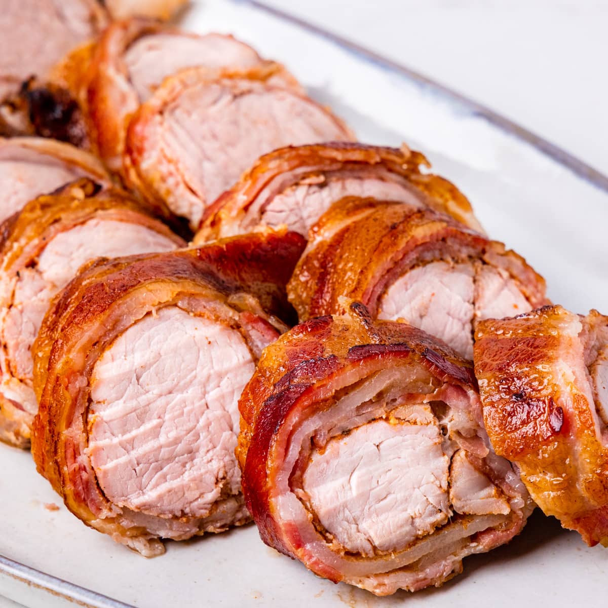 Close up image of sliced bacon wrapped smoked pork tenderloin on a serving platter.