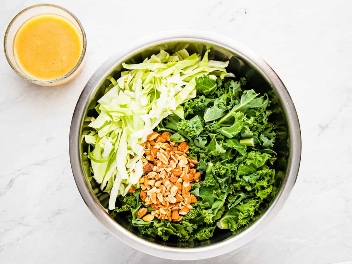 Kale, cabbage, and almonds in a mixing bowl with a small bowl of vinaigrette to the side.