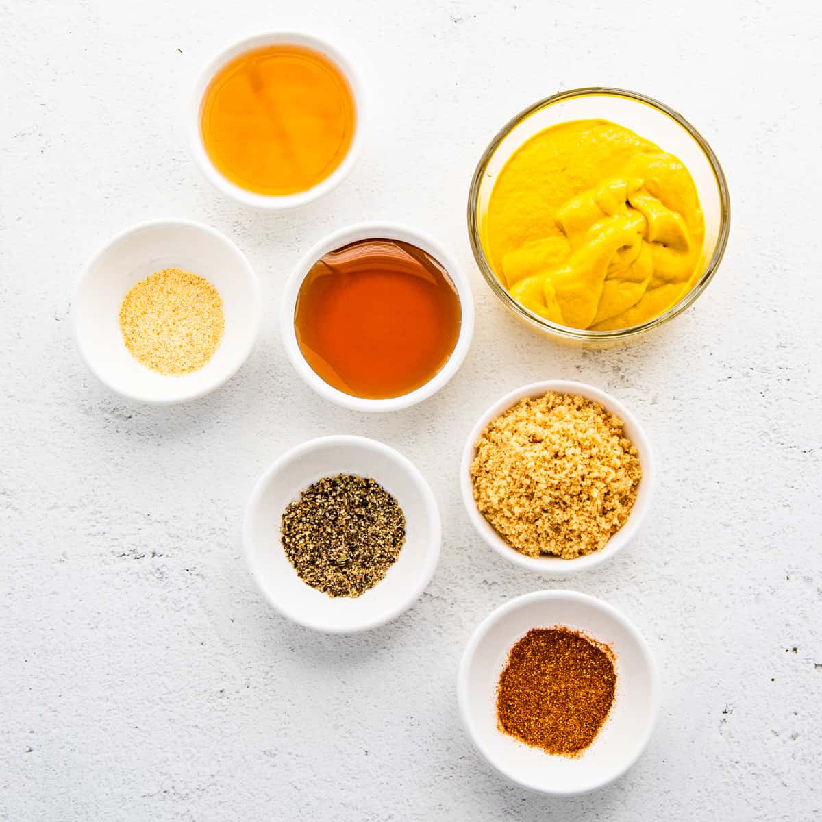 Small bowls of yellow mustard, cider vinegar, honey, garlic powder, black pepper, and cayenne pepper shown set on a counter.