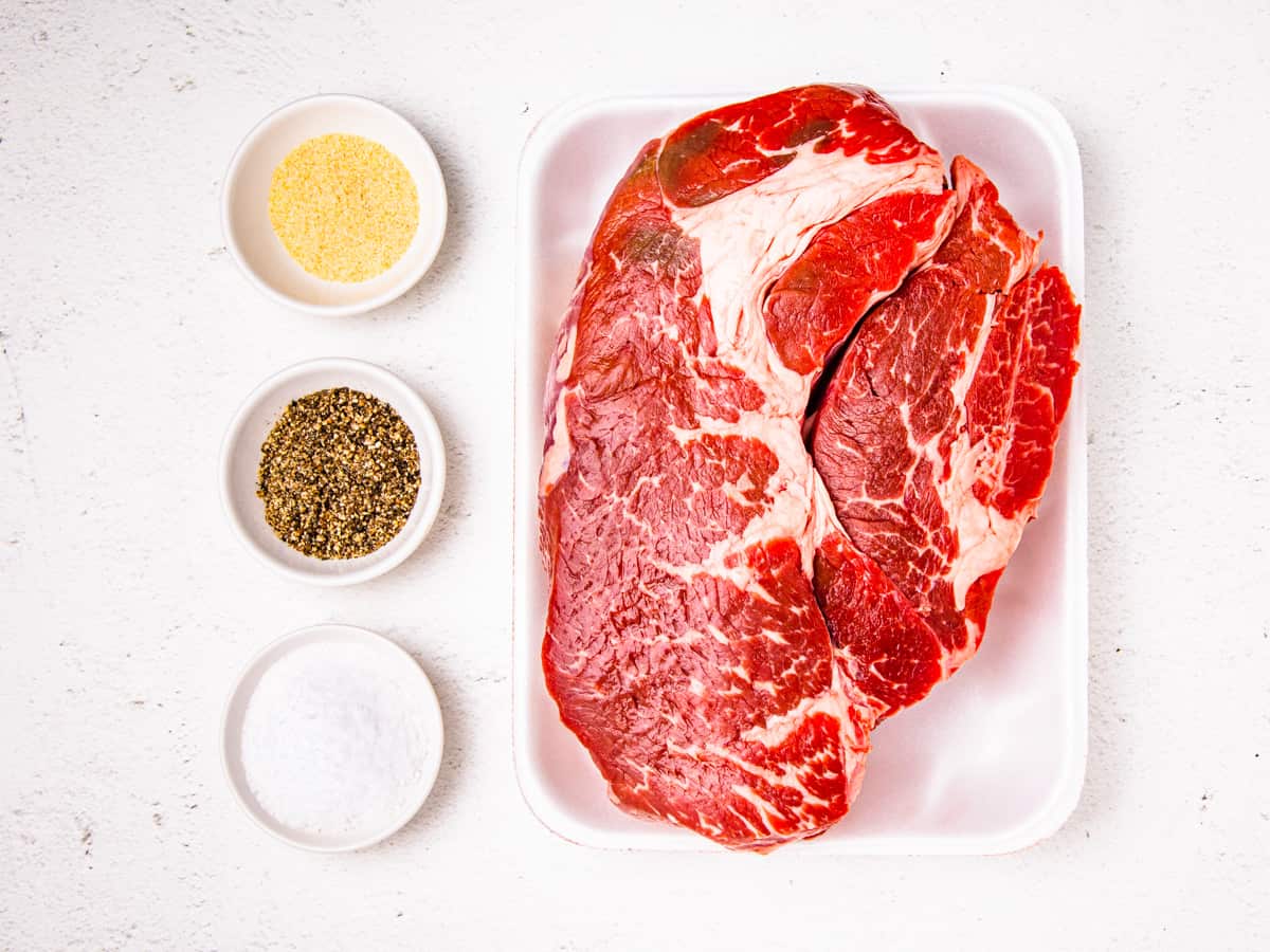 A chuck roast shown on a counter with bowls of salt, pepper, and garlic powder.