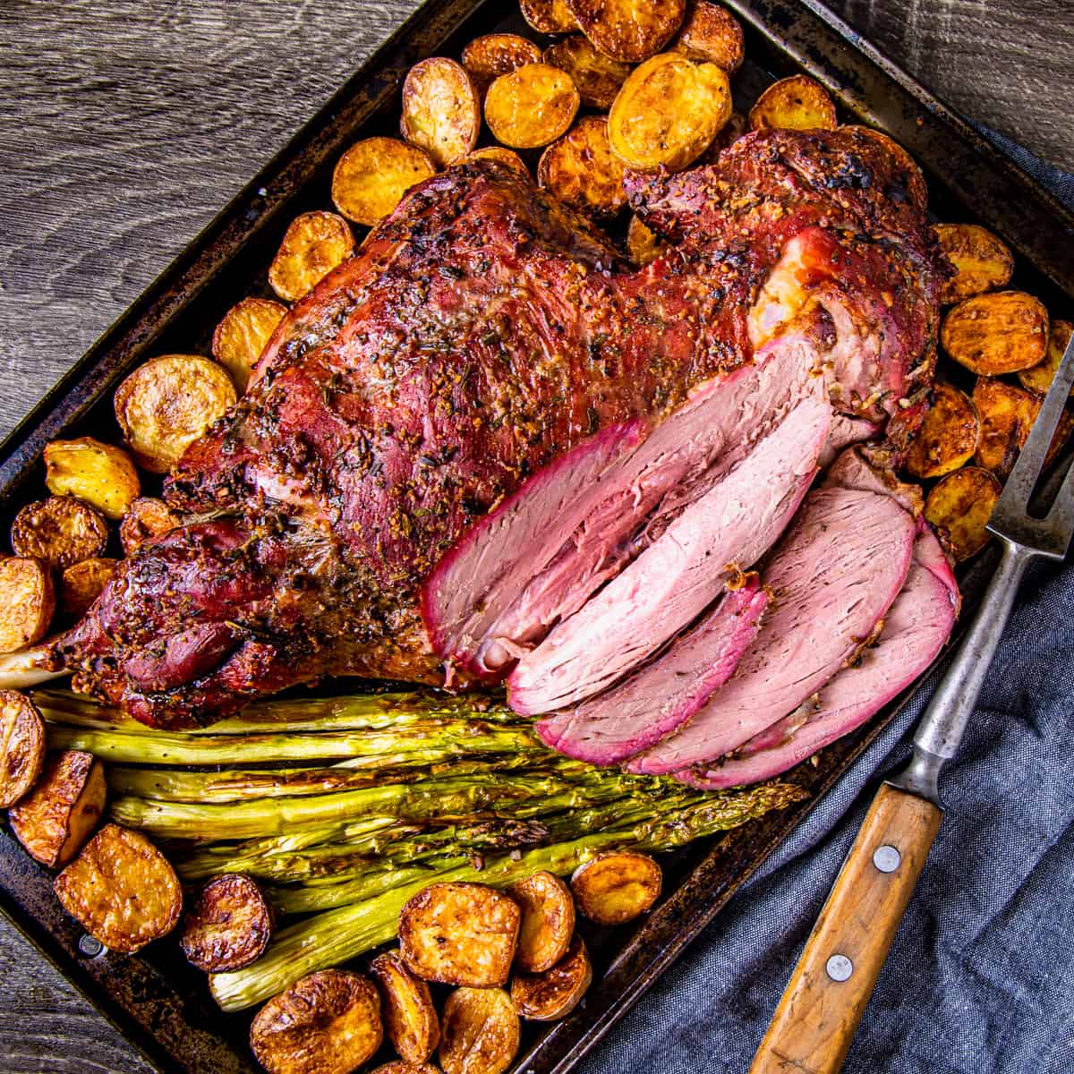 Smoked leg of lamb shown on a sheet pan with roasted potatoes and asparagus. 