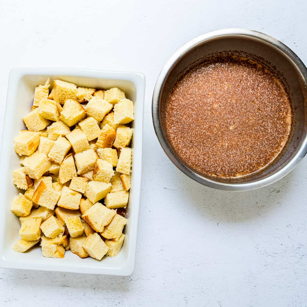 A baking dish full of cubed bread set beside a bowl of custard mixture.