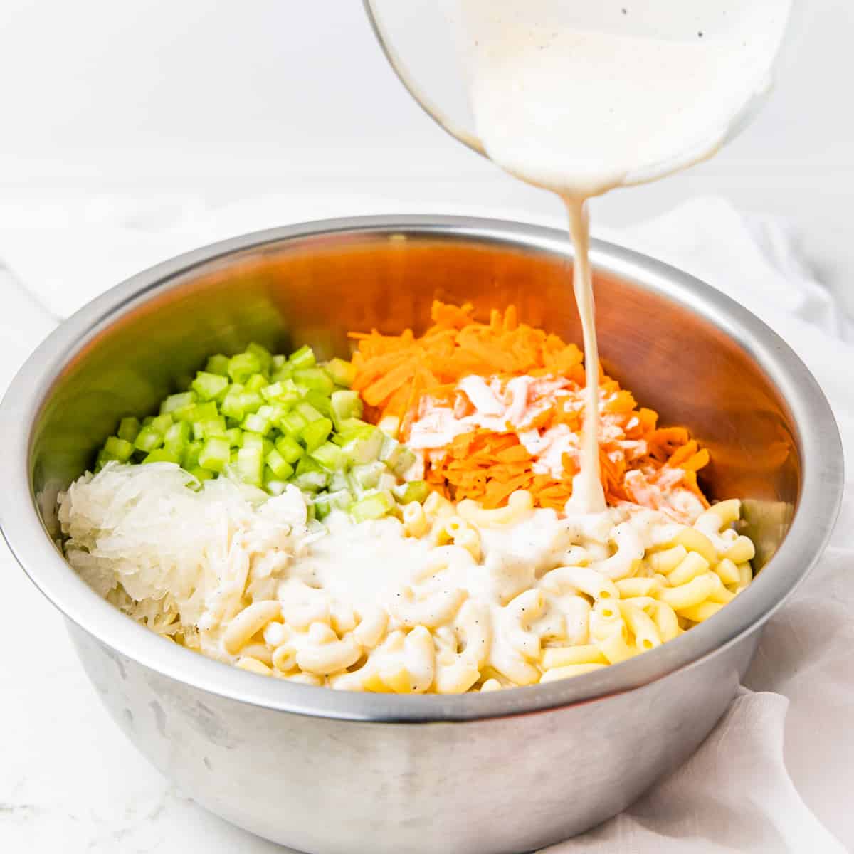 Dressing being drizzled over a large mixing bowl filled with macaroni, carrots, and celery. 