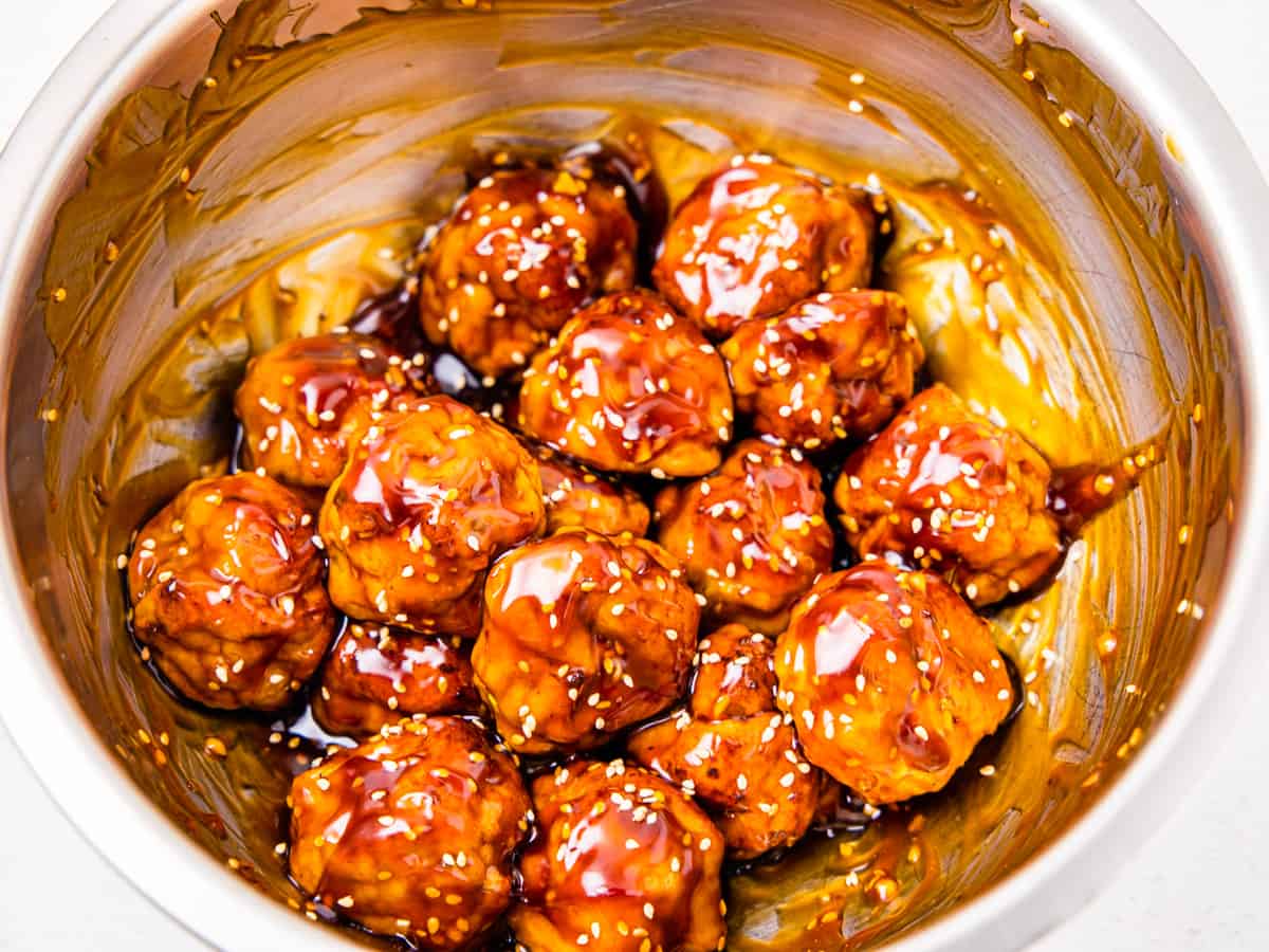 Chicken meatballs are shown in a large mixing bowl being tossed with an Asian sauce. 