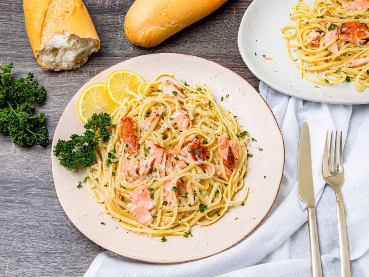 A table setting showing a plate of salmon pasta served with a loaf crusty bread beside a large serving platter of pasta. 