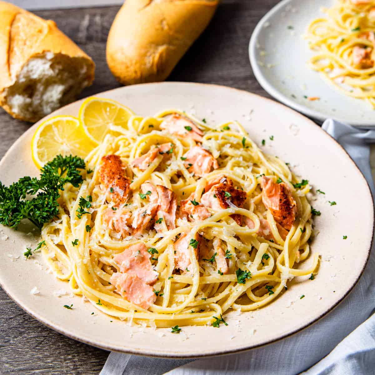 A plate of creamy salmon linguine  shown on a table with a loaf crusty bread and a large serving platter of pasta. 