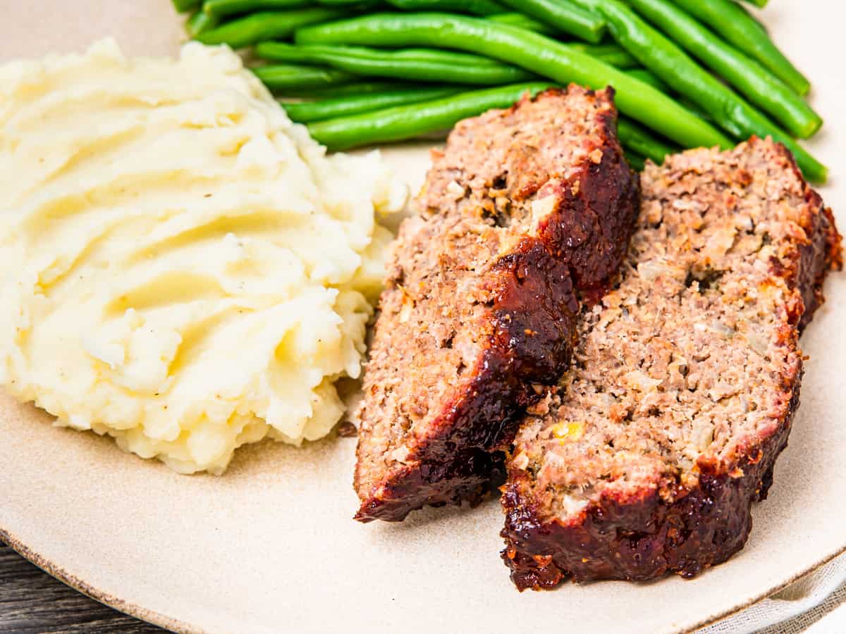 A serving of smoked meatloaf on a plate with mashed potatoes and green beans. 