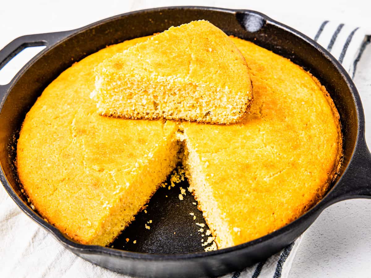 Cornbread in a cast iron skillet with a wedge-shaped slice removed and set on top. 