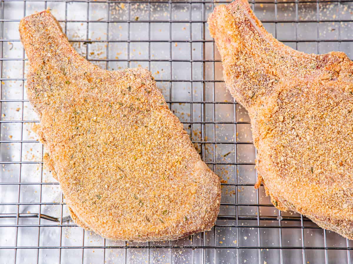 Pork chops coated in a seasoned breadcrumb mixture shown on a baking rack set on a sheet pan prior to baking. 
